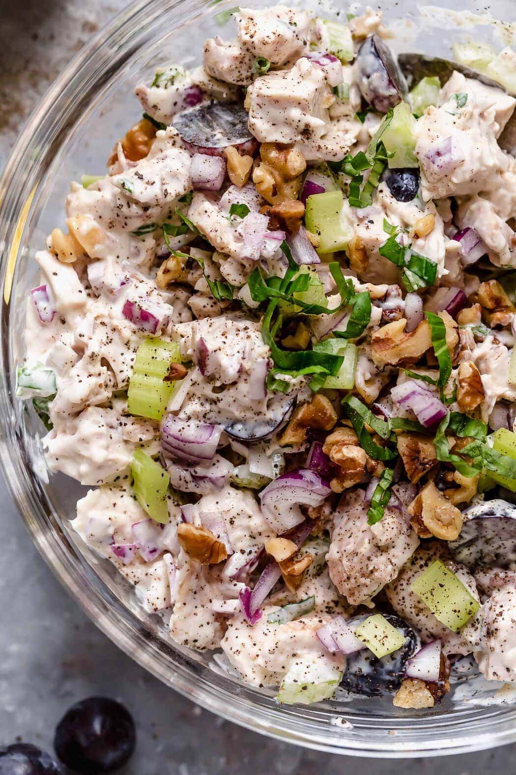 Close up view of Waldorf Chicken Salad in a clear bowl, topped with chopped fresh basil and chopped walnuts. 