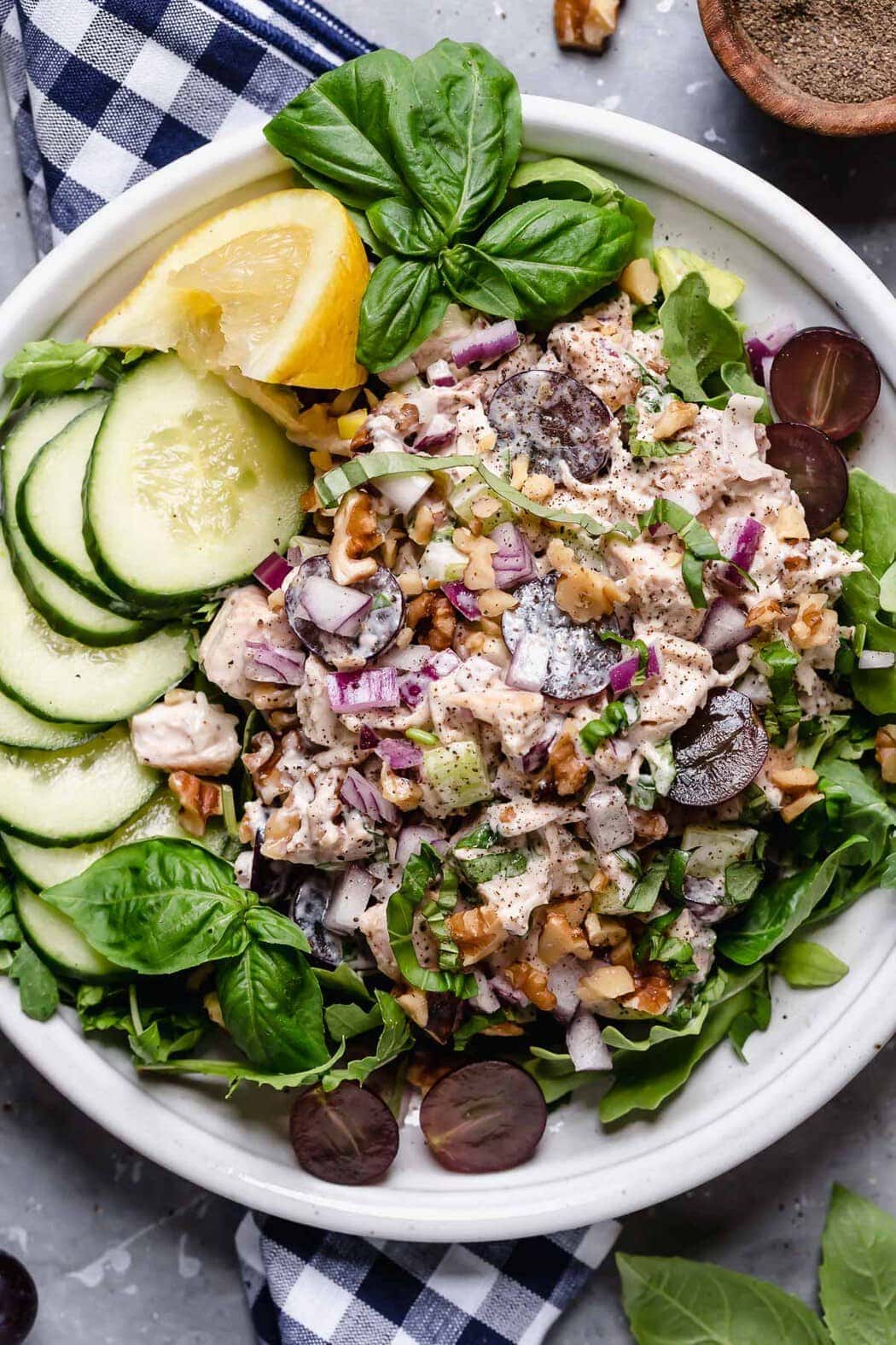 Plate filled with a bed of greens topped with chicken Waldorf salad garnished with cucumbers, grapes and lemon over a checkered napkin