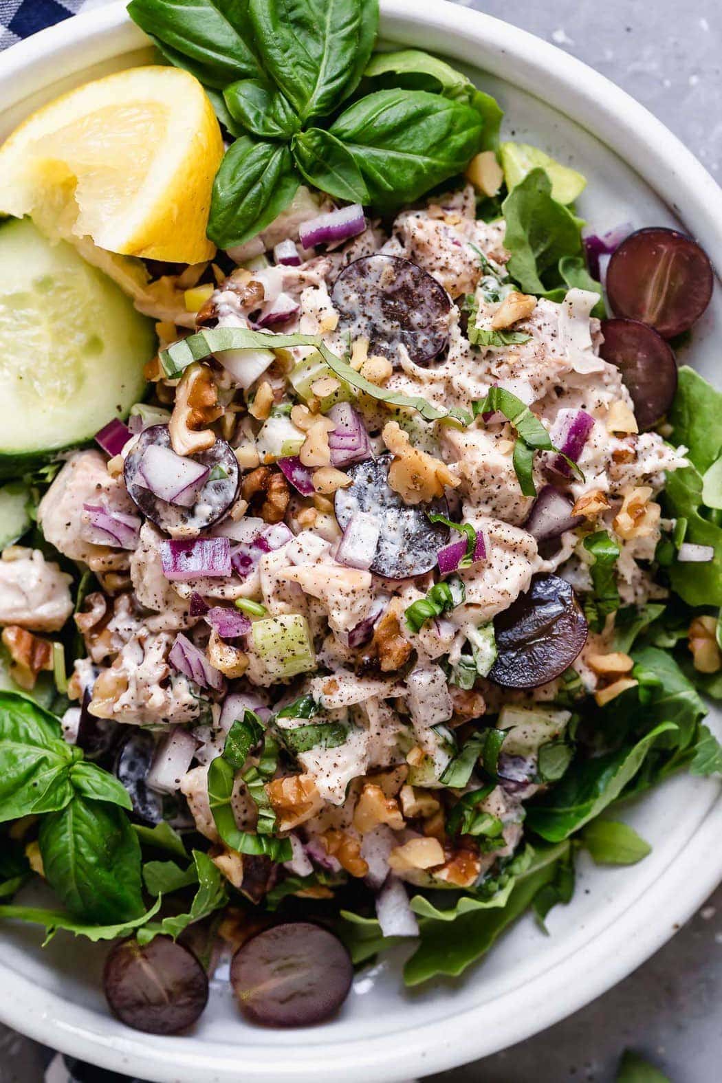 Overhead photo of Classic Waldorf Chicken Salad on a white plate served on top of greens along with cucumber slices, lemon wedge and fresh basil. 