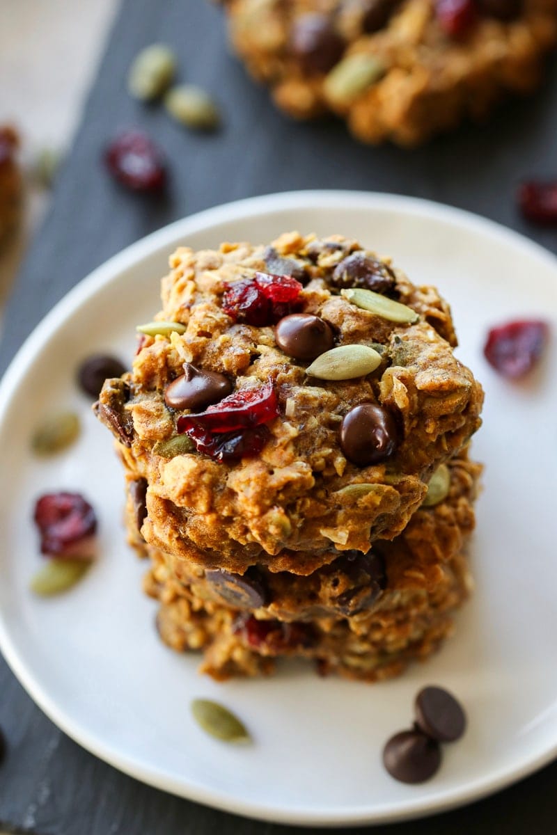 Healthy pumpkin breakfast cookies stacked up on a white plate with chocolate chips and dried cranberries on top.