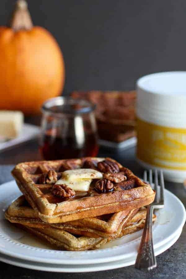 Paleo Pumpkin Spice Protein Waffles on a white plate with a fork resting on the plate and a glass jar of syrup in the background