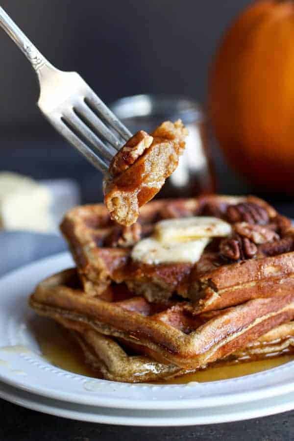 A bite of Paleo Pumpkin Spice Protein Waffles raised towards the camera with a stack of three waffles below it on a white plate