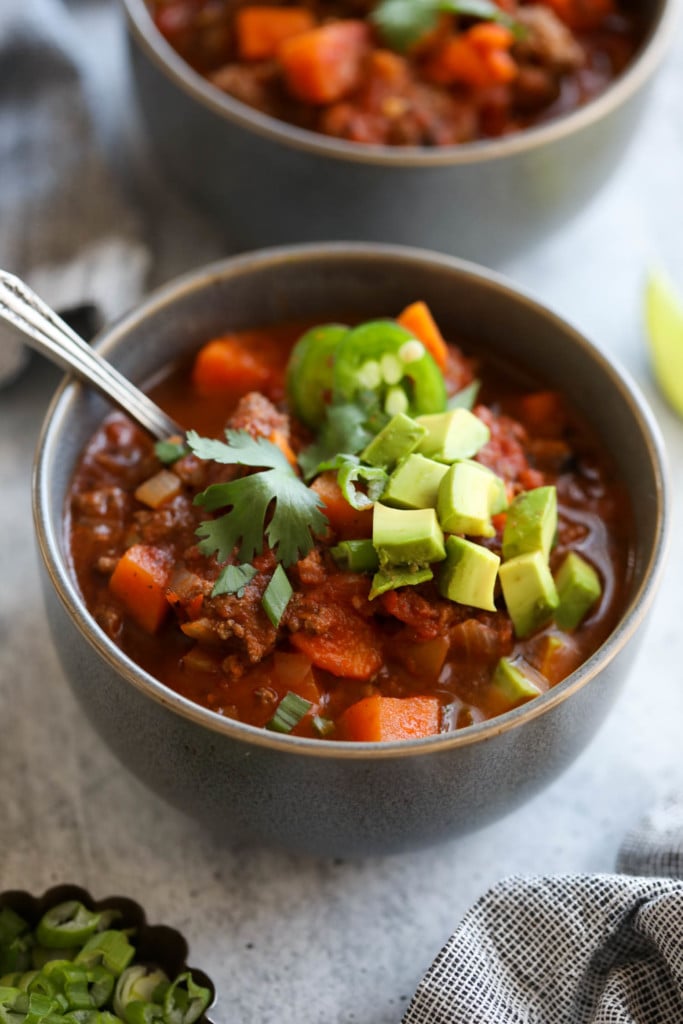 Close up view of slow cooker sweet potato chili with big chunky pieces of sweet potato, tomato, and beef in a bowl served with avocado and fresh cilantro.