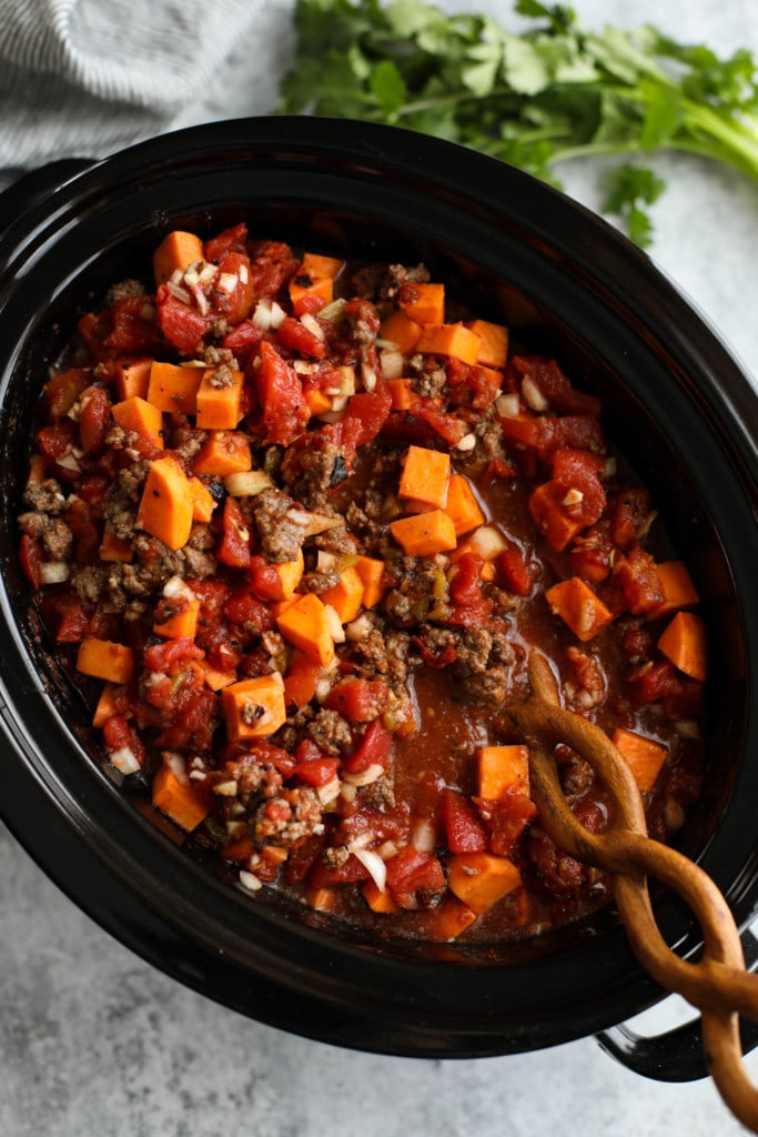 All ingredients for sweet potato chili stirred together with a twisted handle wooden spoon in a black slow cooker.
