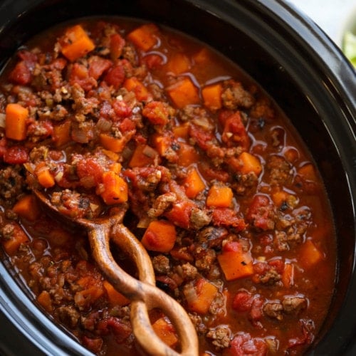 Chunk sweet potato chili being stirred with a twisted handle wooden spoon in a black slow cooker