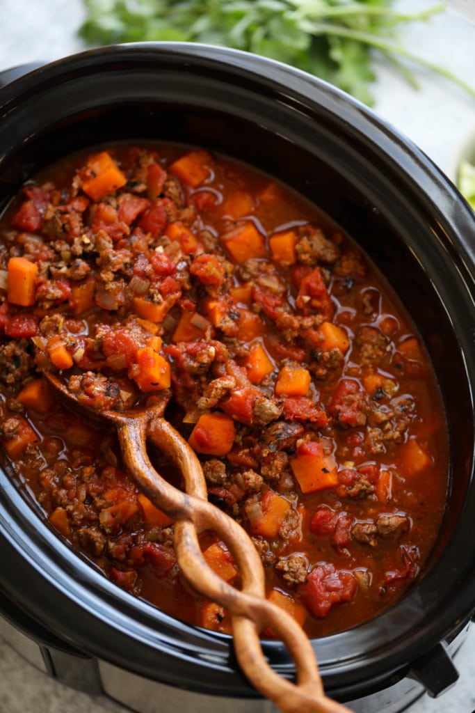 Overhead view of sweet potato chili in a black slow cooker with a twisted handle wooden spoon stirring the chili.