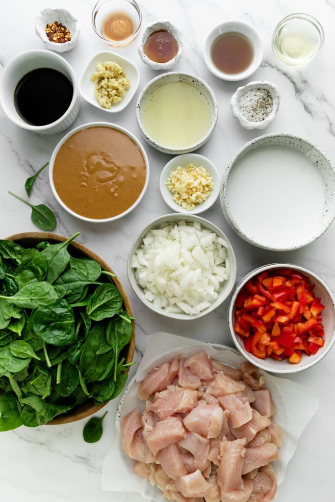 All ingredients for Thai-inspired peanut chicken in bowls of different sized arranged together on a marble countertop.