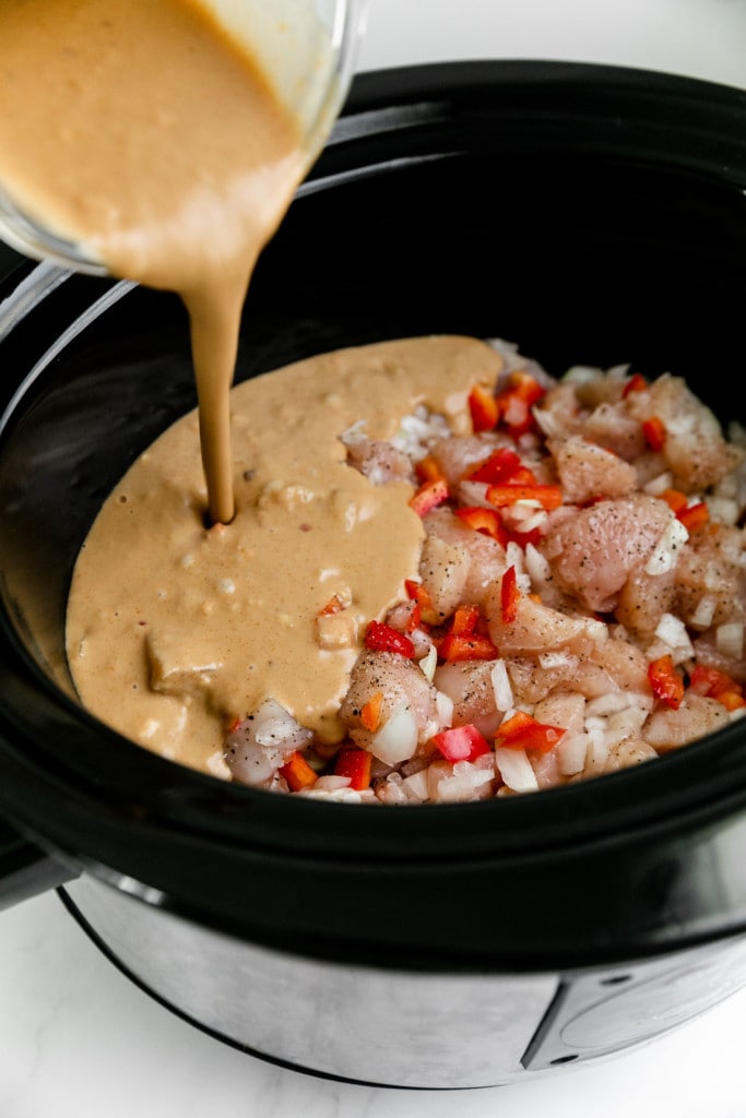 Homemade peanut sauce being poured into a slow cooker filled with raw chicken and diced red pepper. 