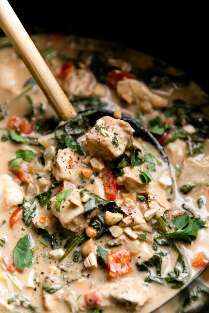 Close up view of creamy Thai peanut chicken in a slow cooker with a black spoon stirring it. 