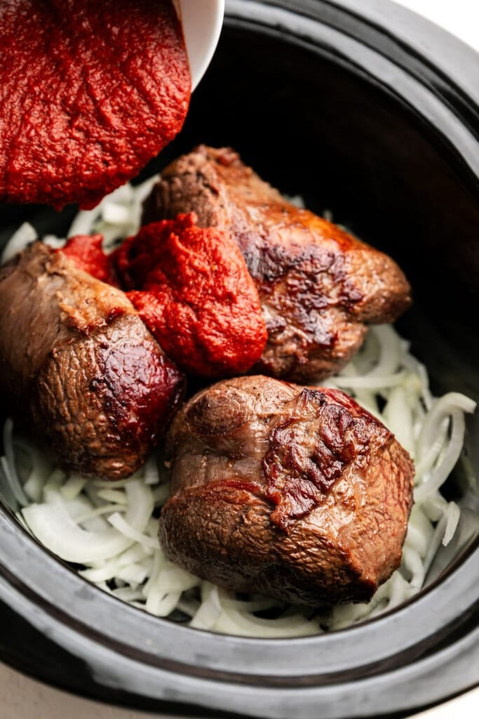 BBQ sauce being poured over browned beef in black slow cooker