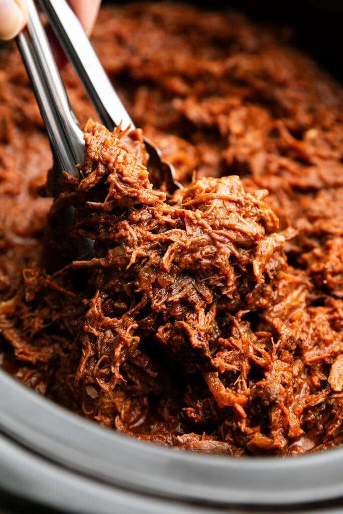 Shredded bbq beef being served with tongs from black slow cooker