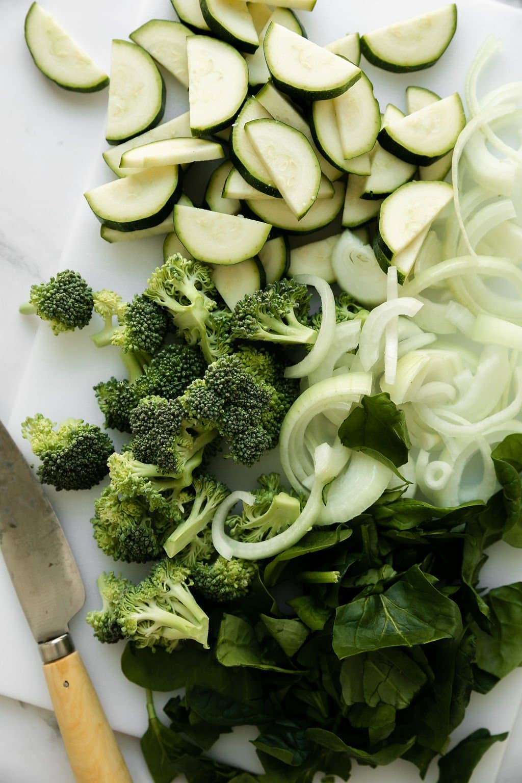 Chopped zucchini, broccoli, onion, and spinach on a white cutting board with a knife on the side to add to 5-Ingredient Baked Ziti as a veggie-loaded option.