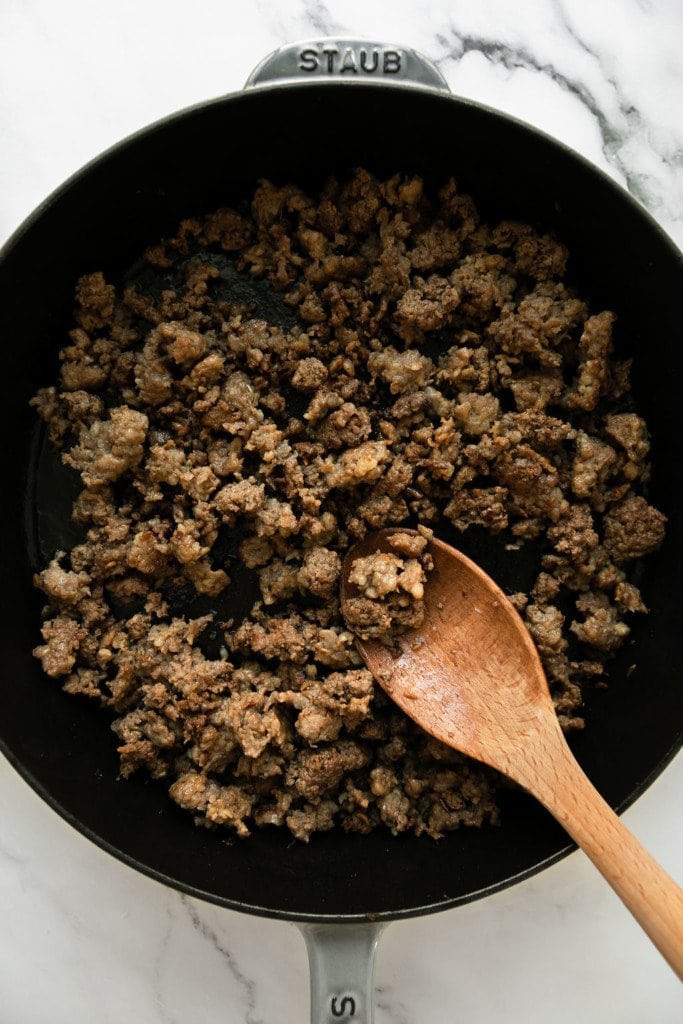 Overhead view of browned ground beef and ground pork in a skillet with a wooden spoon stirring it.