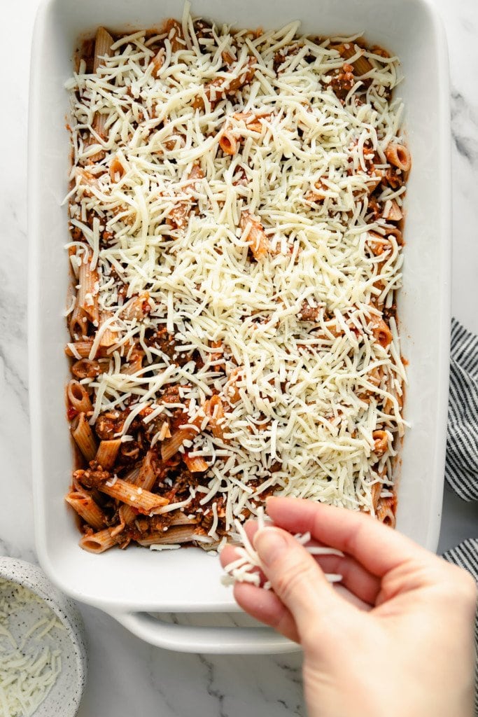 Overhead view of uncooked baked ziti in a white baking dish with a hand sprinkling shredded mozzarella cheese on top.