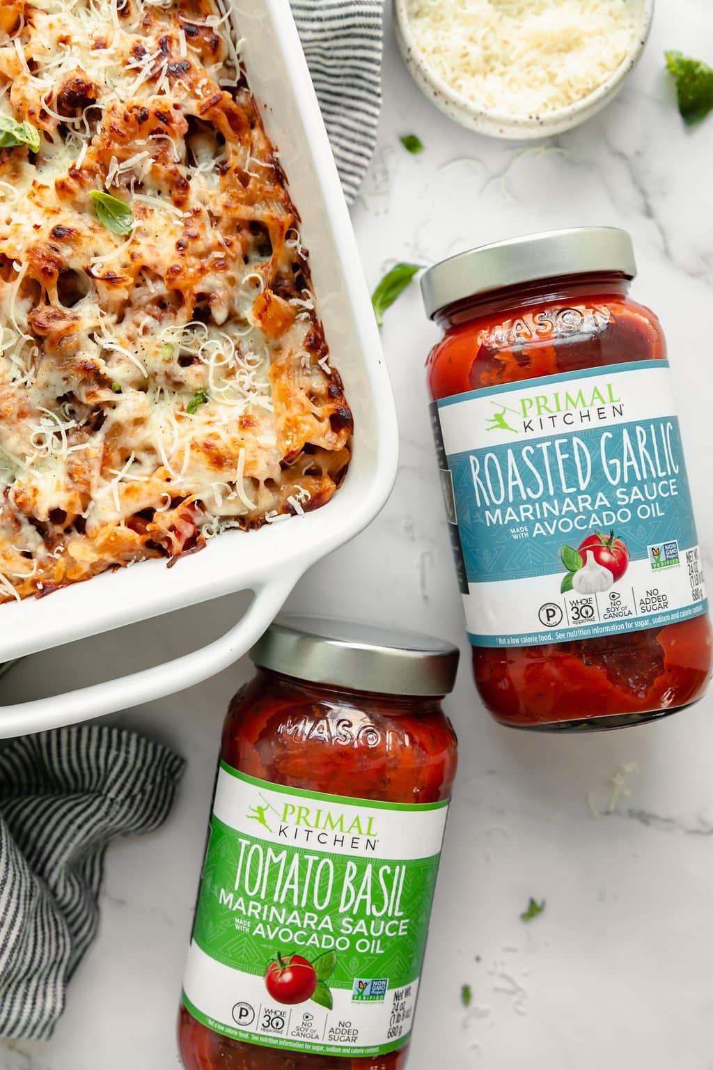 A baked ziti in a white casserole dish with melted mozzarella cheese with two jars of Primal Kitchen marinara sauce next to the dish.