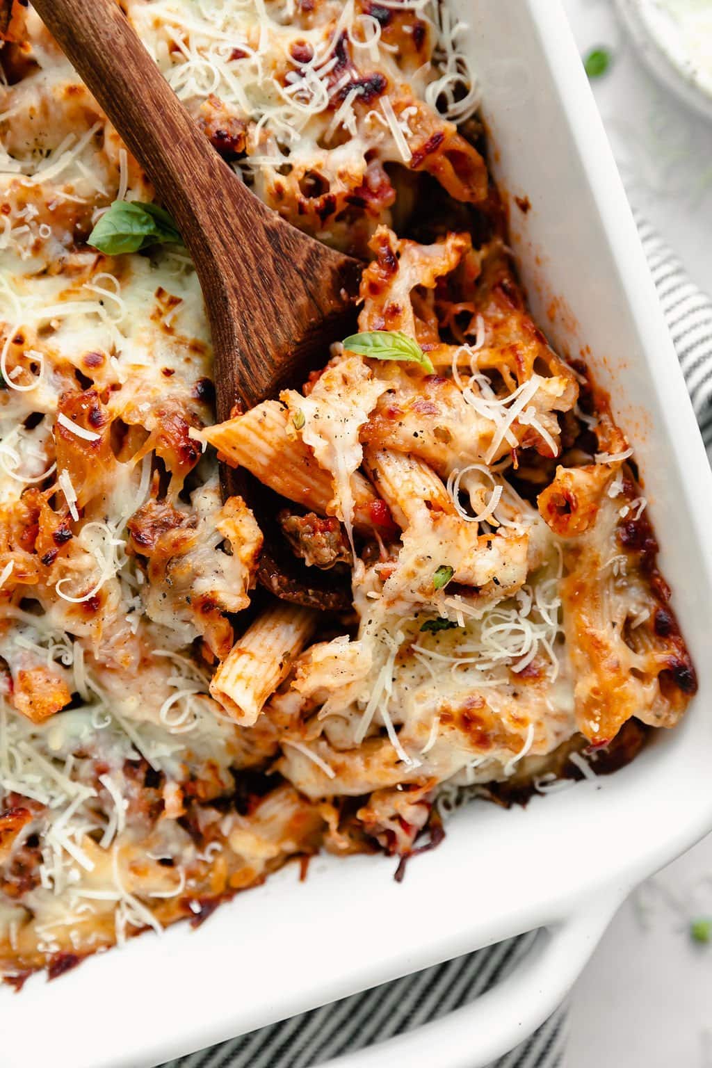 A wooden spoon scooping out a serving of 5-ingredient baked ziti with a melted mozzarella cheese from a white baking dish.
