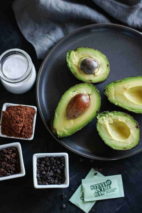 Avocados, cocoa powder, chocolate chips, coconut milk and packets of stevia on a plate. 