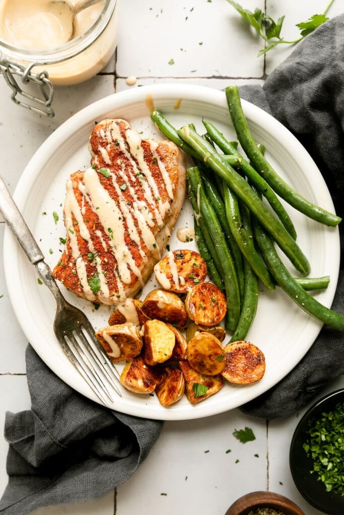 White plate with serving of baked pork chop with honey mustard drizzled over top side of roasted potato halves and green beans.