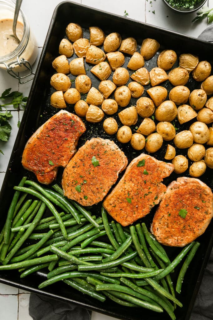 Overhead view black baking sheet filled with potato halves, pork chops, and green beans.