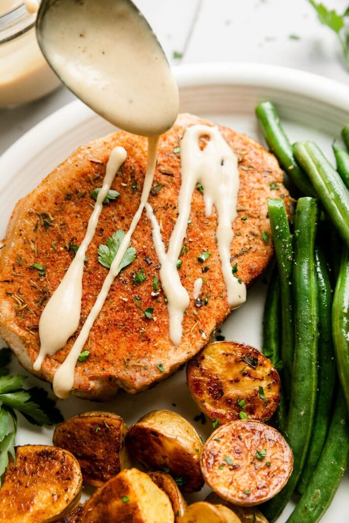 Honey mustard being drizzled over baked pork chop on white plate with side of green beans and potatoes