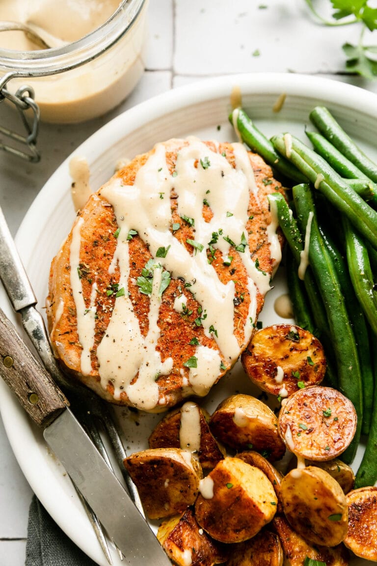 Close up view pork chop with honey mustard sauce drizzled over top, on a white plate roasted potato halves and green beans.