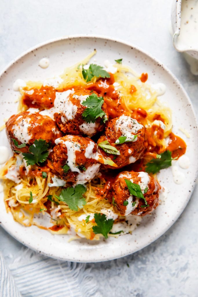 Buffalo Chicken Meatballs served over spaghetti squash, topped with a drizzle of ranch and garnished with cilantro and green onion. Served on a white plate. 