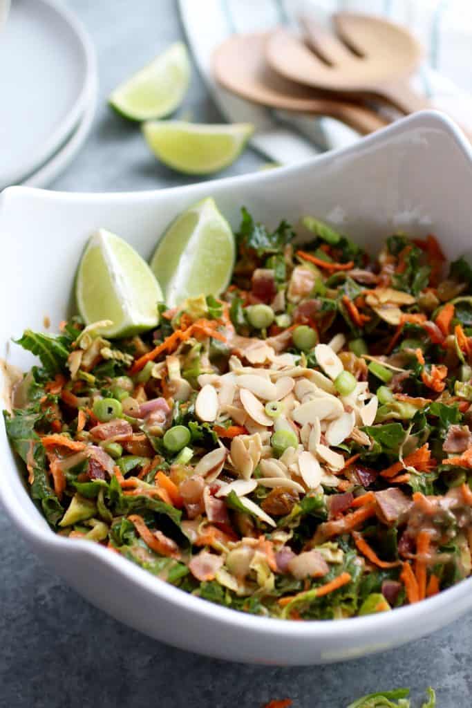 BBQ Ranch chopped salad with Brussels sprouts and kale in a white serving bowl topped with sliced almonds and lime wedges.