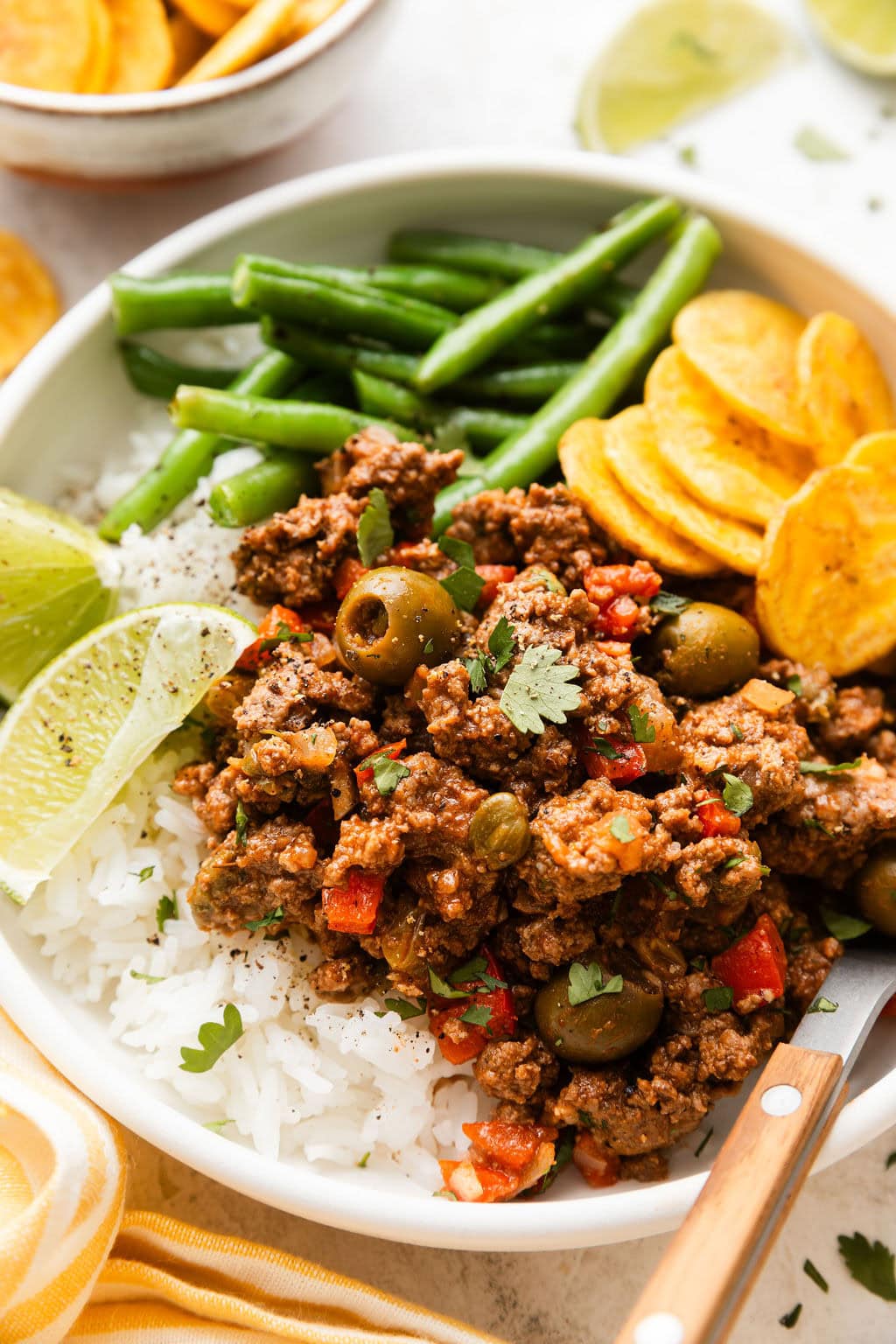 Close up view of a plate of Stovetop Picadillo on top of rice next to plantain chips and green beans topped with fresh cilantro. 