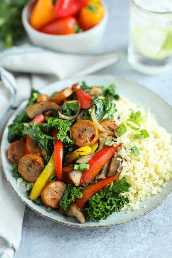 Bowl of Grilled Chicken Sausage and Veggies over Cauliflower rice on a table with a glass of ice water. 