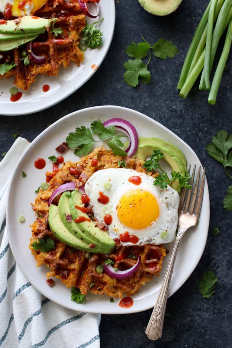 Overhead view of a plate filled with a sweet potato bacon waffle topped with a fried egg and sliced avocado.