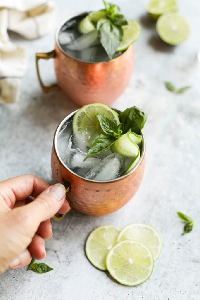 A hand holding a copper cup handle filled with cucumber basil Moscow Mule garnished with lime and cucumber