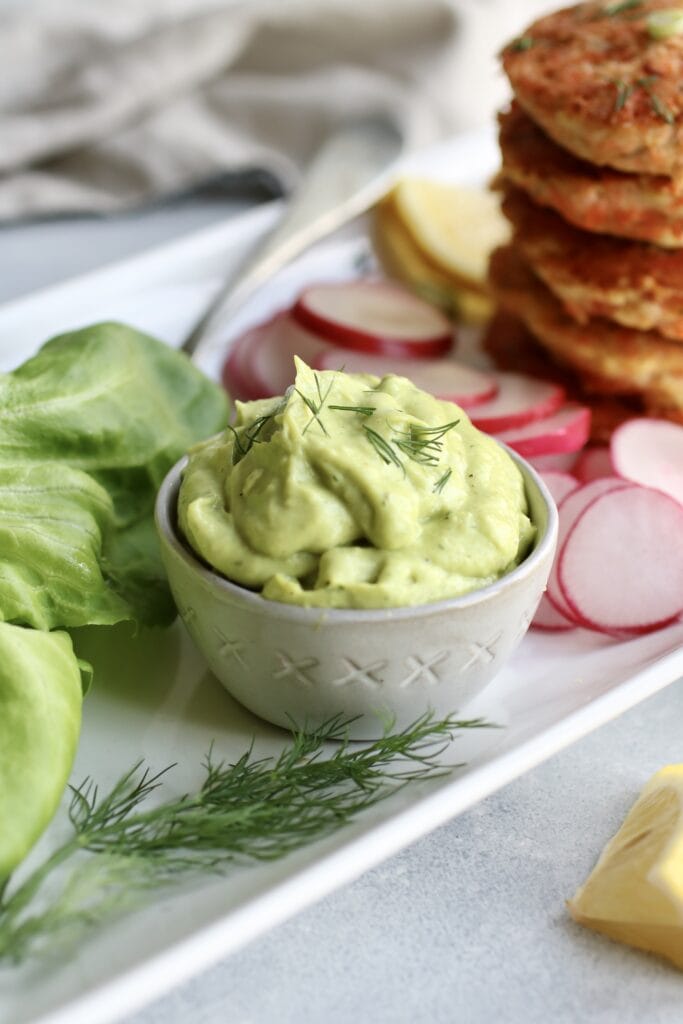 Creamy avocado sauce in small white bowl with stack of salmon burgers to the side