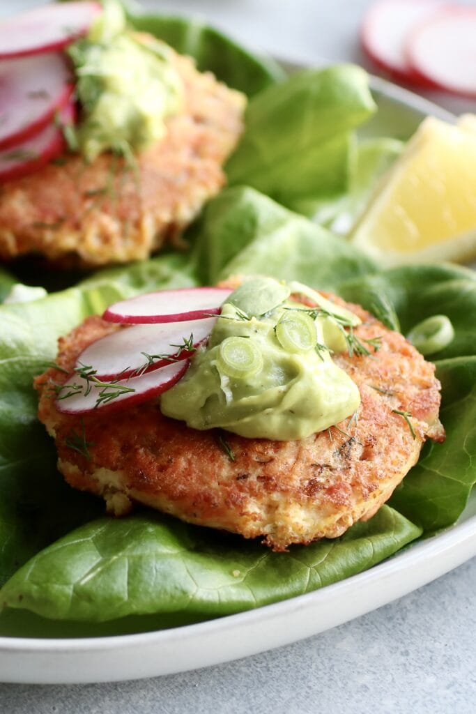 Close up view pan fried salmon burger on lettuce leaves topped with avocado sauce and sliced radishes