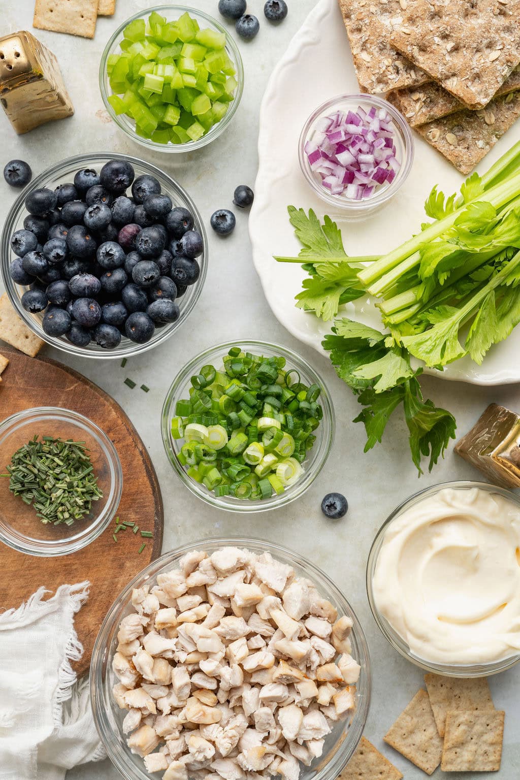 Overhead view of a variety of ingredients for Blueberry Chicken Salad in different sized bowls on a white marble countertop.