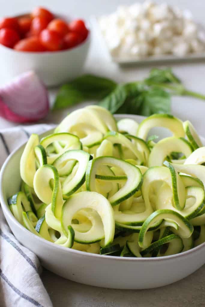Spiralized zucchini in white bowl with fresh veggies in background