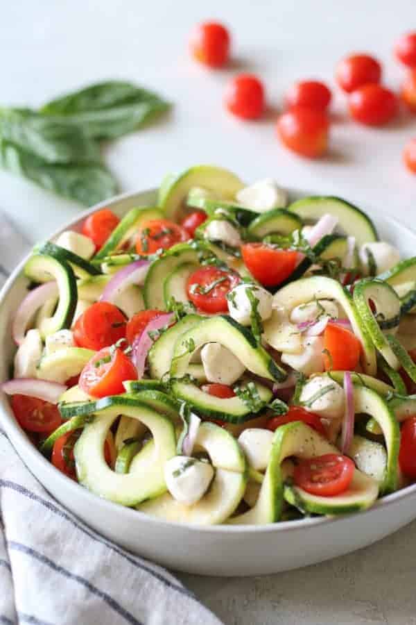 Caprese Zucchini Salad with Balsamic Vinaigrette in a white bowl with cherry tomatoes and herbs in the background