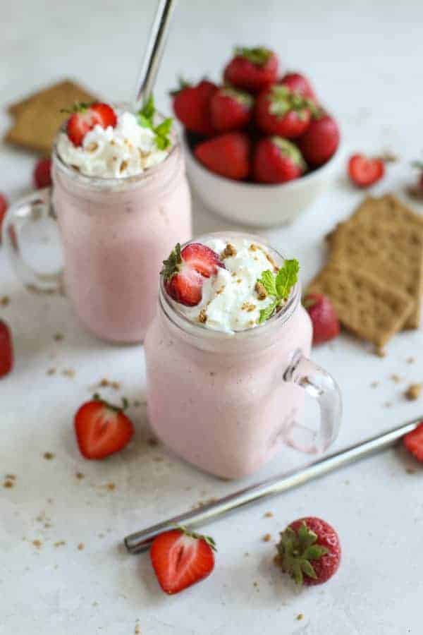 Two Strawberry Cheesecake Smoothies in glasses, topped with whipped cream and graham cracker crumbs sprinkled all around. 