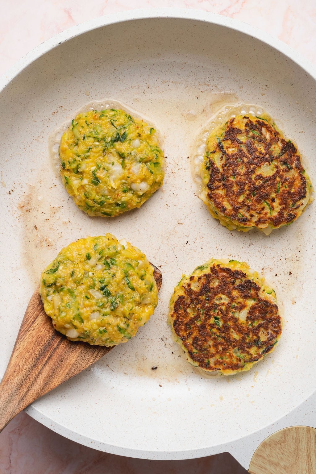 A wooden spatula flipping four zucchini fritters in a skillet one at a time.