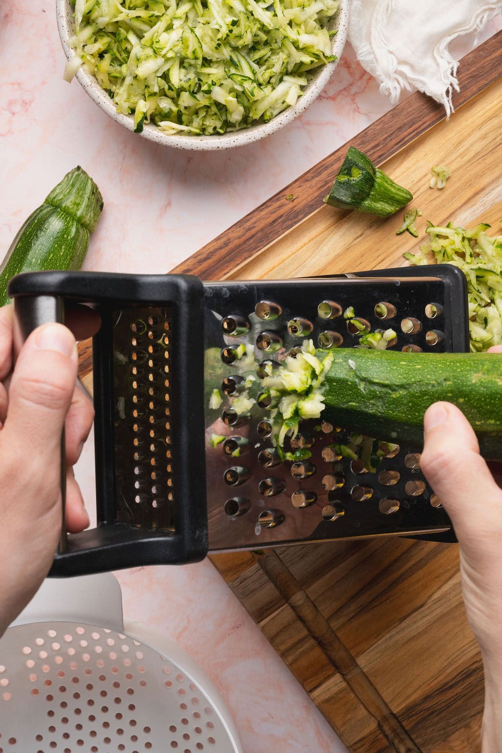 Hands using a box grater to shred raw zucchini.