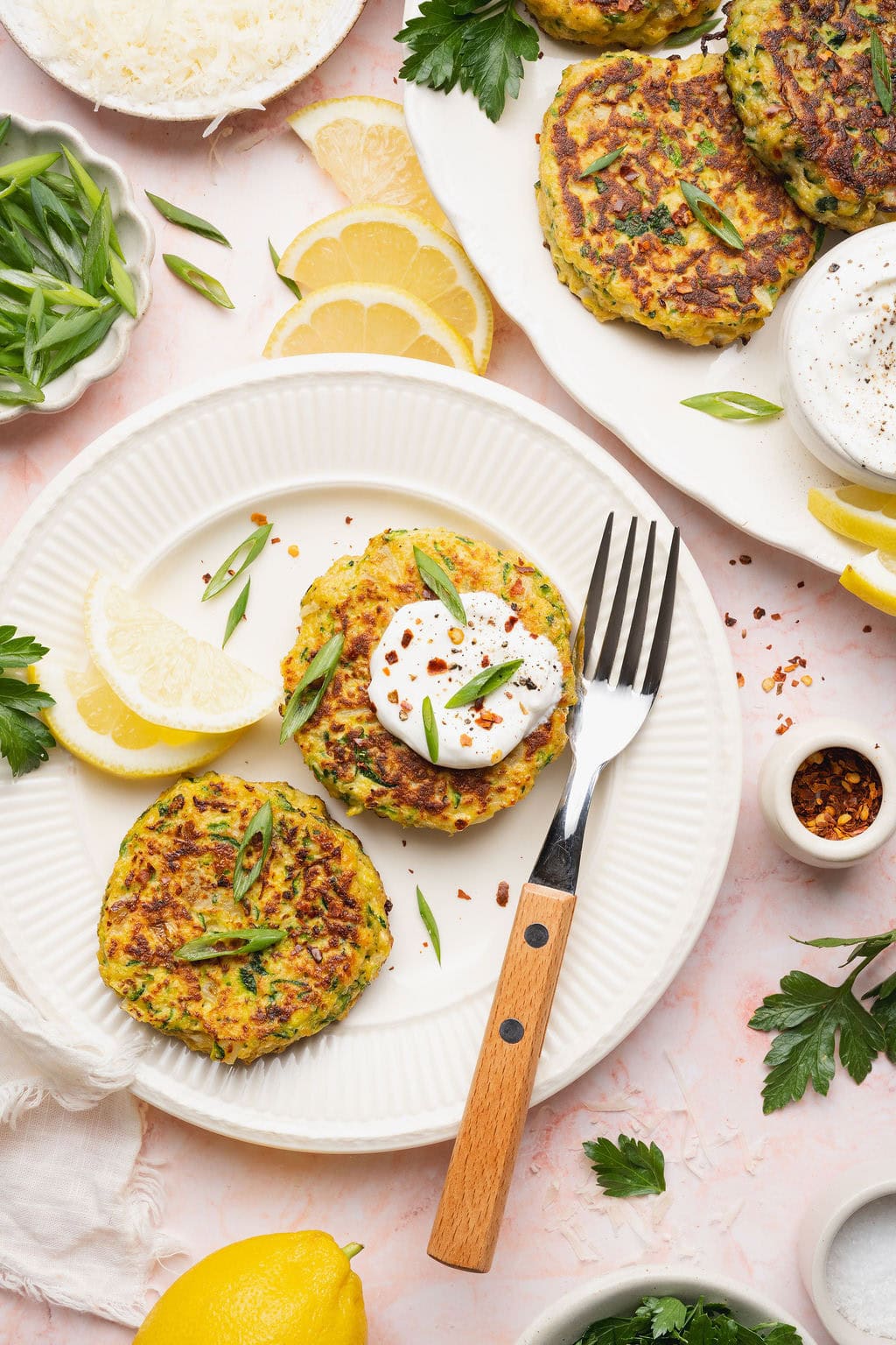 Two zucchini fritters on a plate next to a fork, two lemon wedges on the side and a dollop of mayonnaise on top of one.