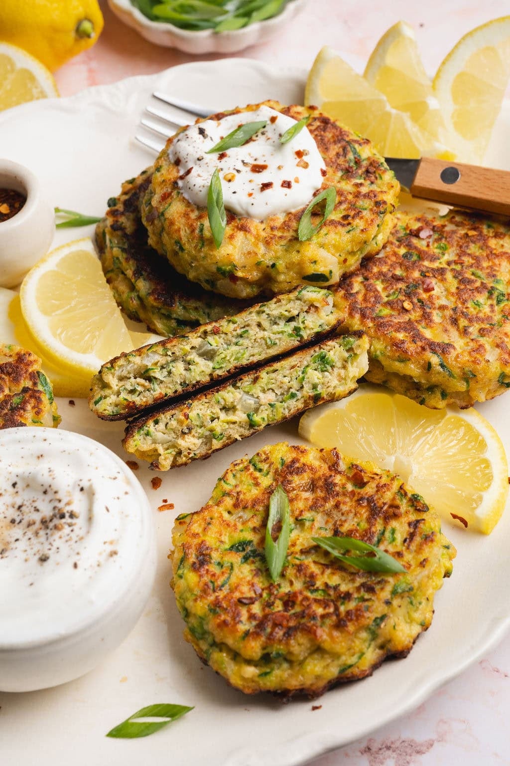A platter of zucchini fritters with a small bowl of mayo, one fritter split in half, lemon slices around them.