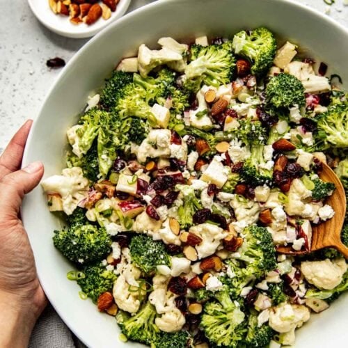 Large serving bowl filled with apple broccoli cauliflower salad tossed in lemony dressing being held by two hands.