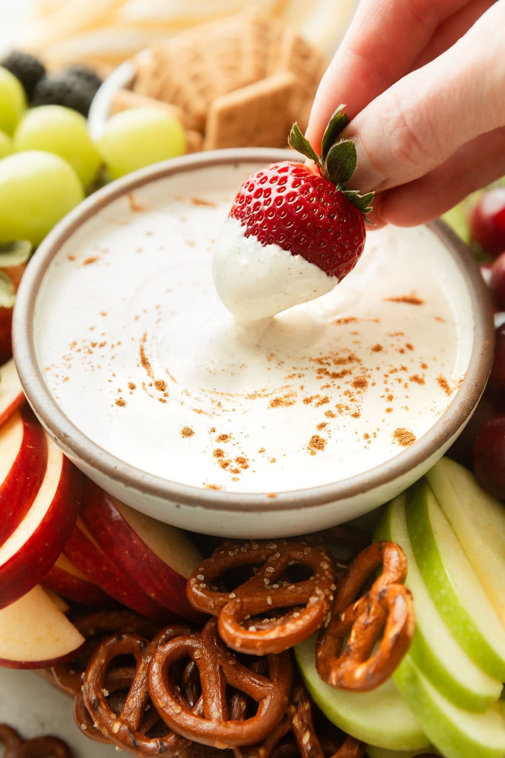 Close up view of a hand dipping a fresh strawberry into a fluffy white fruit dip sprinkled with cinnamon.