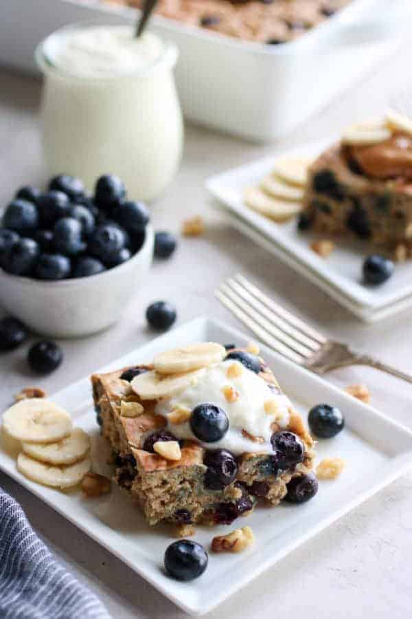 A square piece of baked oatmeal on white square plate surrounded by small bowl of blueberries, a glass cup filled with yogurt and a glimpse of the full pan as well as another plated piece