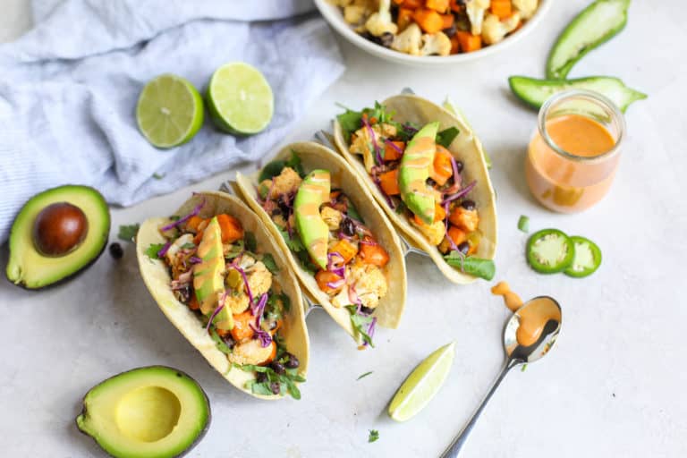 Three cauliflower sweet potato tacos in taco holder topped with avocado and dressing.