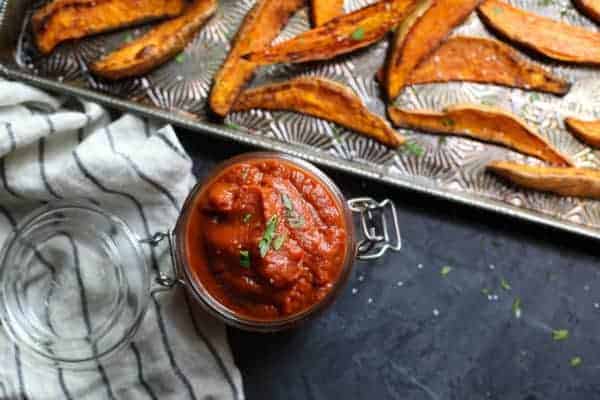 Overhead view of jar of BBQ sauce with roasted sweet potato wedges on a baking pan next to it. 