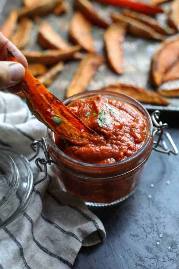 Hand dipping a roasted sweet potato wedge into a jar of barbecue sauce. 