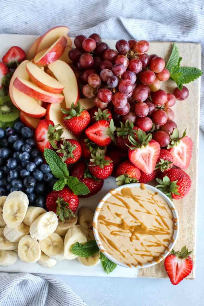 Overhead view wooden flat tray filled with fresh cut fruit; bowl of peanut butter yogurt fruit dip nestled into fruit