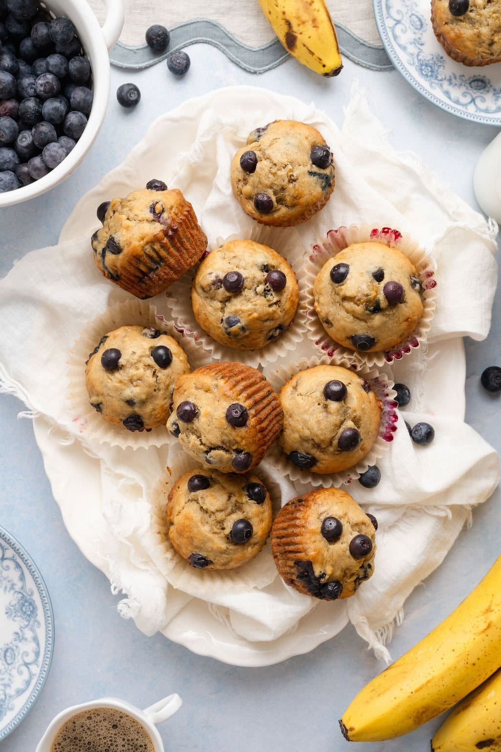Overhead view of a batch of freshly baked Banana Blueberry Muffins on a white dish towel. 