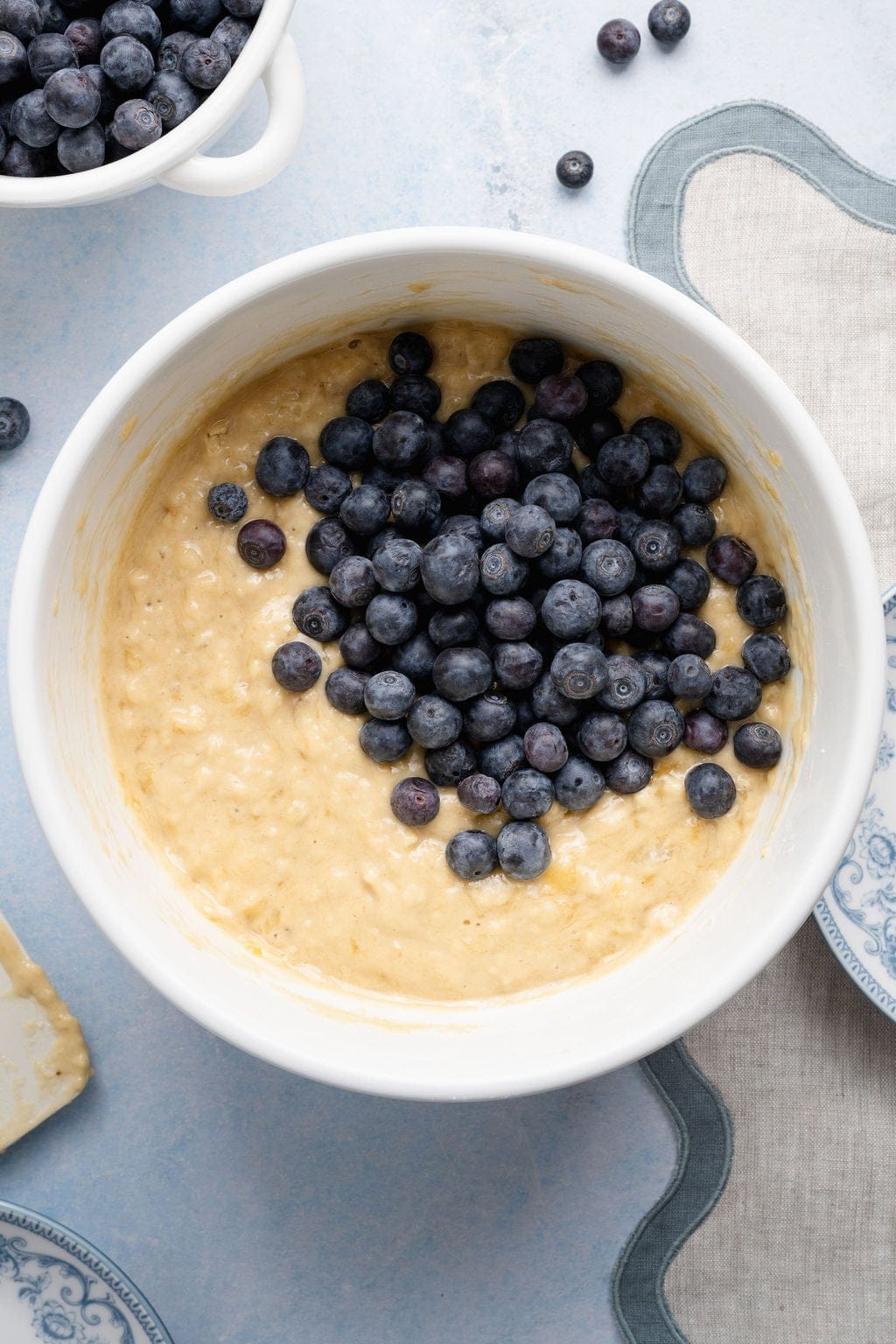 Overhead view of a white bowl filled with Banana Blueberry Muffin dough topped with fresh blueberries ready for stirring. 